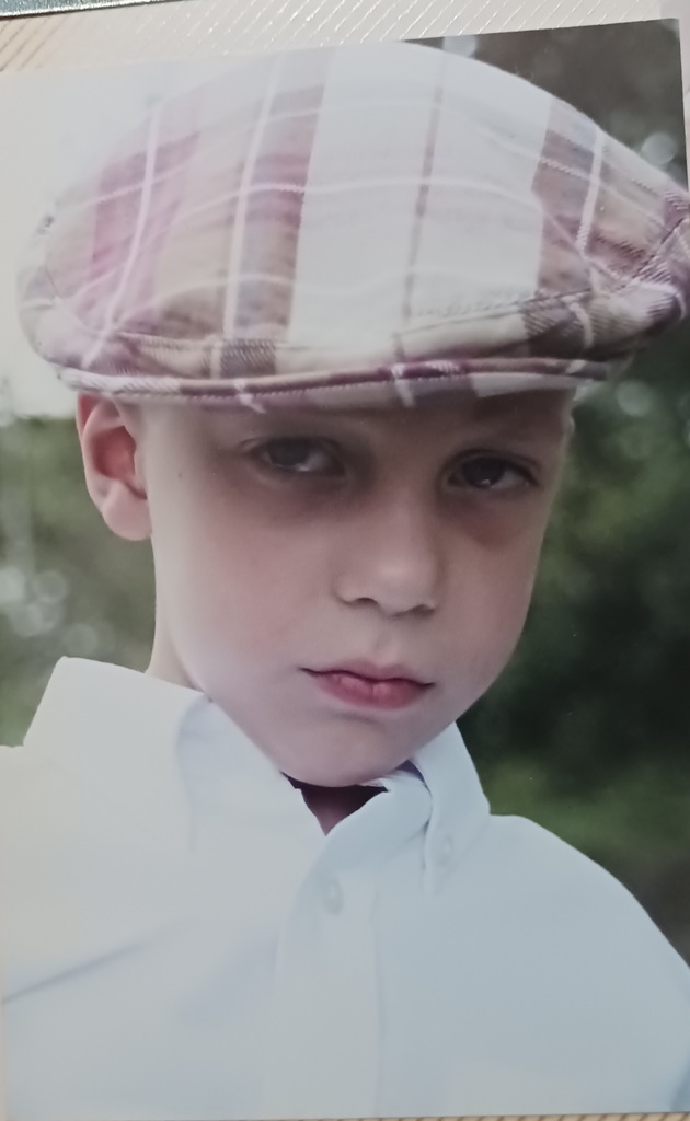 A young boy in a plaid flat cap and a white button-down shirt, looking toward the camera.