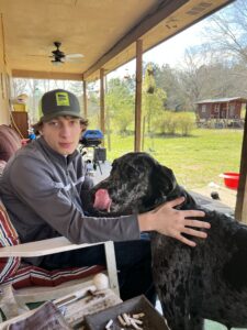 Teenage boy on a covered porch in a cap and jacket, hand resting on a large dog beside him.