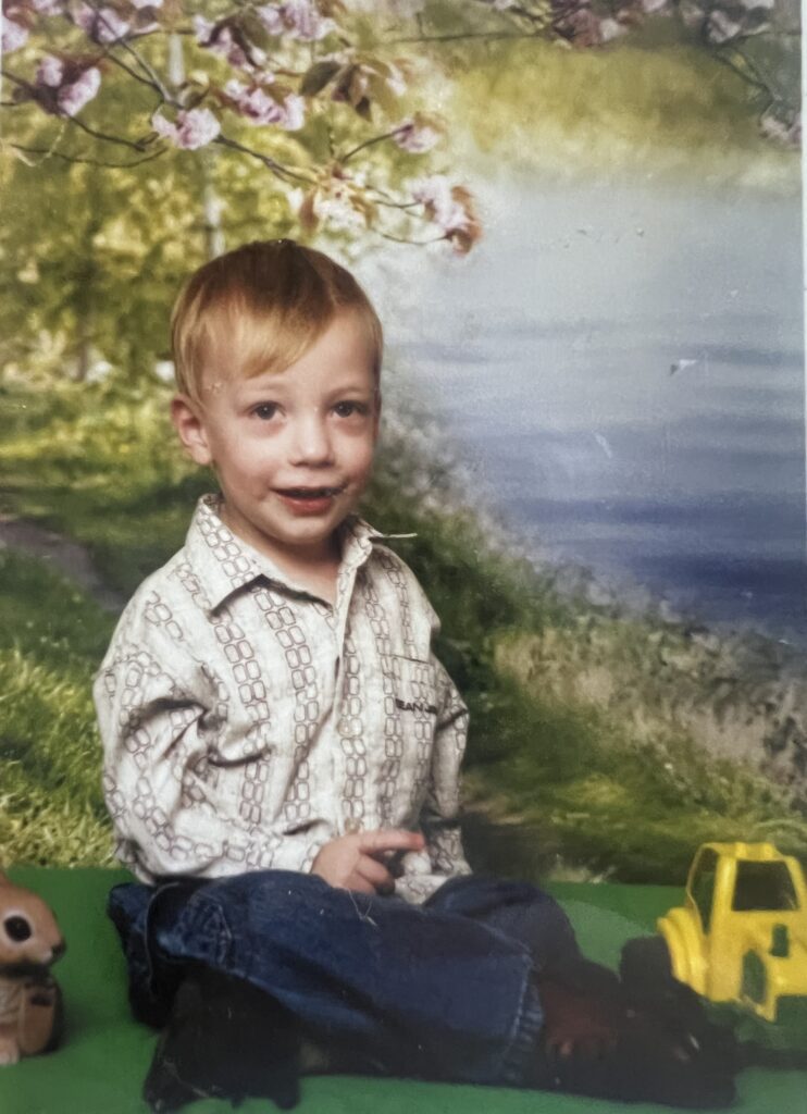 Old studio portrait: a toddler seated on grass in front of a painted lake backdrop, a yellow toy truck beside him.