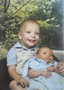 Old studio portrait: a toddler in denim overalls holding a newborn on his lap.
