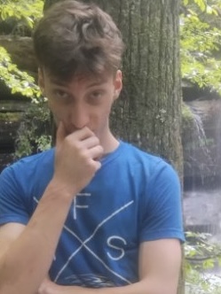 Young man in a blue t-shirt standing by a tree with a waterfall behind him.
