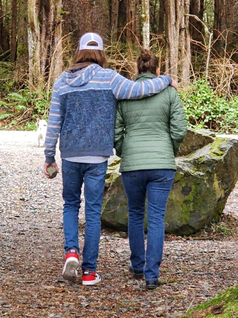 Two people walking away down a gravel trail through the woods, one with an arm around the other's shoulder.